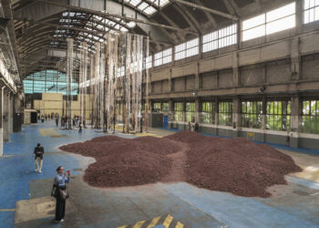 18e Biennale d'art contemporain de Lyon, Les Grandes Locos. Premier plan : Lara Almarcegui, La Montagne de feu, 2026. Schiste rouge. Courtesy de l’artiste. Second plan : Joël Andrianomearisoa, Tady Mitady Tadidy,2026. Fibre et textiles divers, acier, peinture. Impression sur bâche PVC, techniques mixtes. Courtesy de l’artiste et de la Galerie Almine Rech. © Blandine Soulage